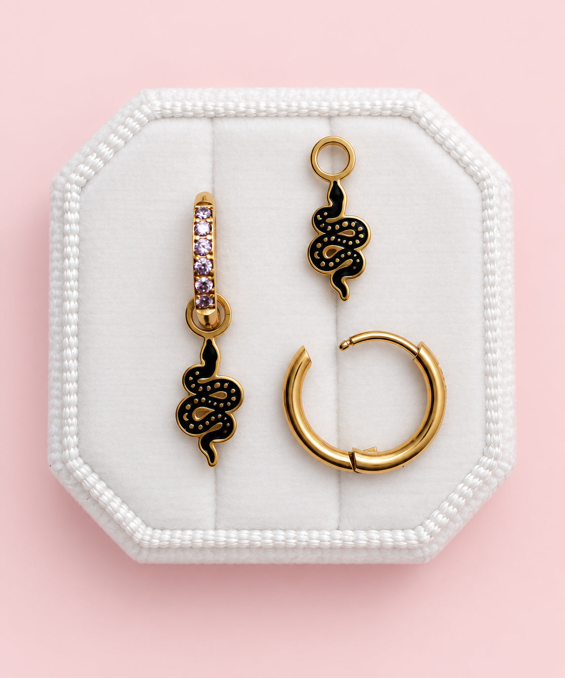 Three gold-toned earrings displayed on a white octagonal jewelry box against a soft pink background. The set includes two snake-shaped drop earrings with black enamel detailing, one with small purple stones, and one large gold hoop earring.