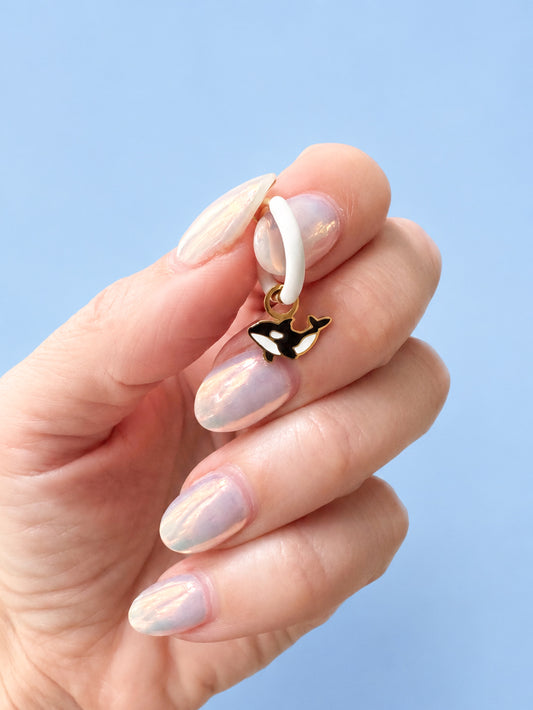 Hand holding a small white hoop with an orca whale charm against a light blue background