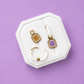 A set of three cute kawaii earrings displayed on a white octagonal velvet jewelry stand against a solid lavender-purple background. The set includes white huggie hoops and peanut butter and jelly shaped charms