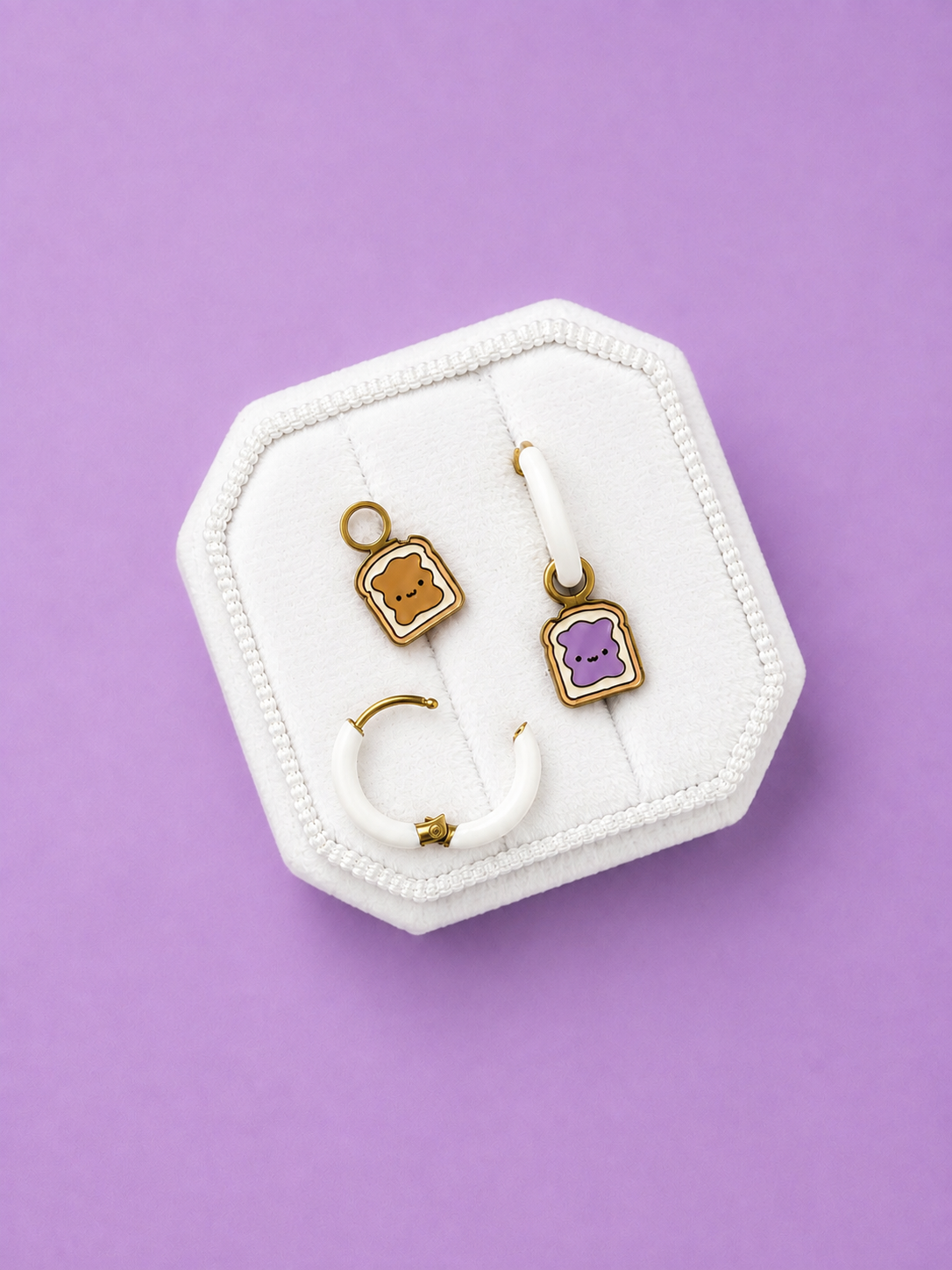 A set of three cute kawaii earrings displayed on a white octagonal velvet jewelry stand against a solid lavender-purple background. The set includes white huggie hoops and peanut butter and jelly shaped charms