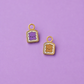 Two small enamel charms shaped like toast slices with cute faces, one purple and one tan, on a solid lavender background.