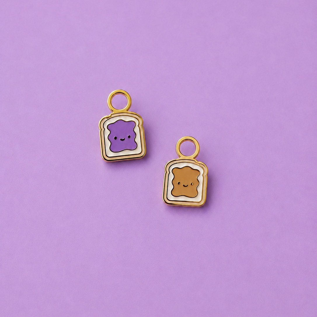 Two small enamel charms shaped like toast slices with cute faces, one purple and one tan, on a solid lavender background.