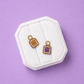 Two small enamel charms shaped like toast pieces with cute faces, displayed on a white octagonal jewelry stand against a solid lavender background. One charm is tan/brown and the other is purple, both with gold-tone rings.