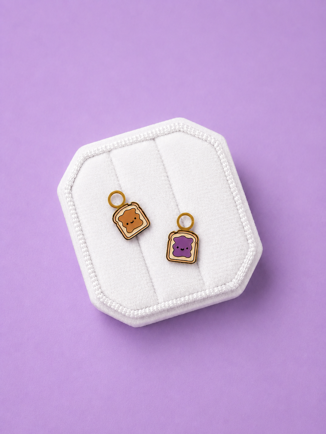 Two small enamel charms shaped like toast pieces with cute faces, displayed on a white octagonal jewelry stand against a solid lavender background. One charm is tan/brown and the other is purple, both with gold-tone rings.