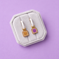 A pair of gold-toned hoop earrings with small enamel charms shaped like peanut butter and jelly slices, displayed on a light gray jewelry stand against a solid lavender background.