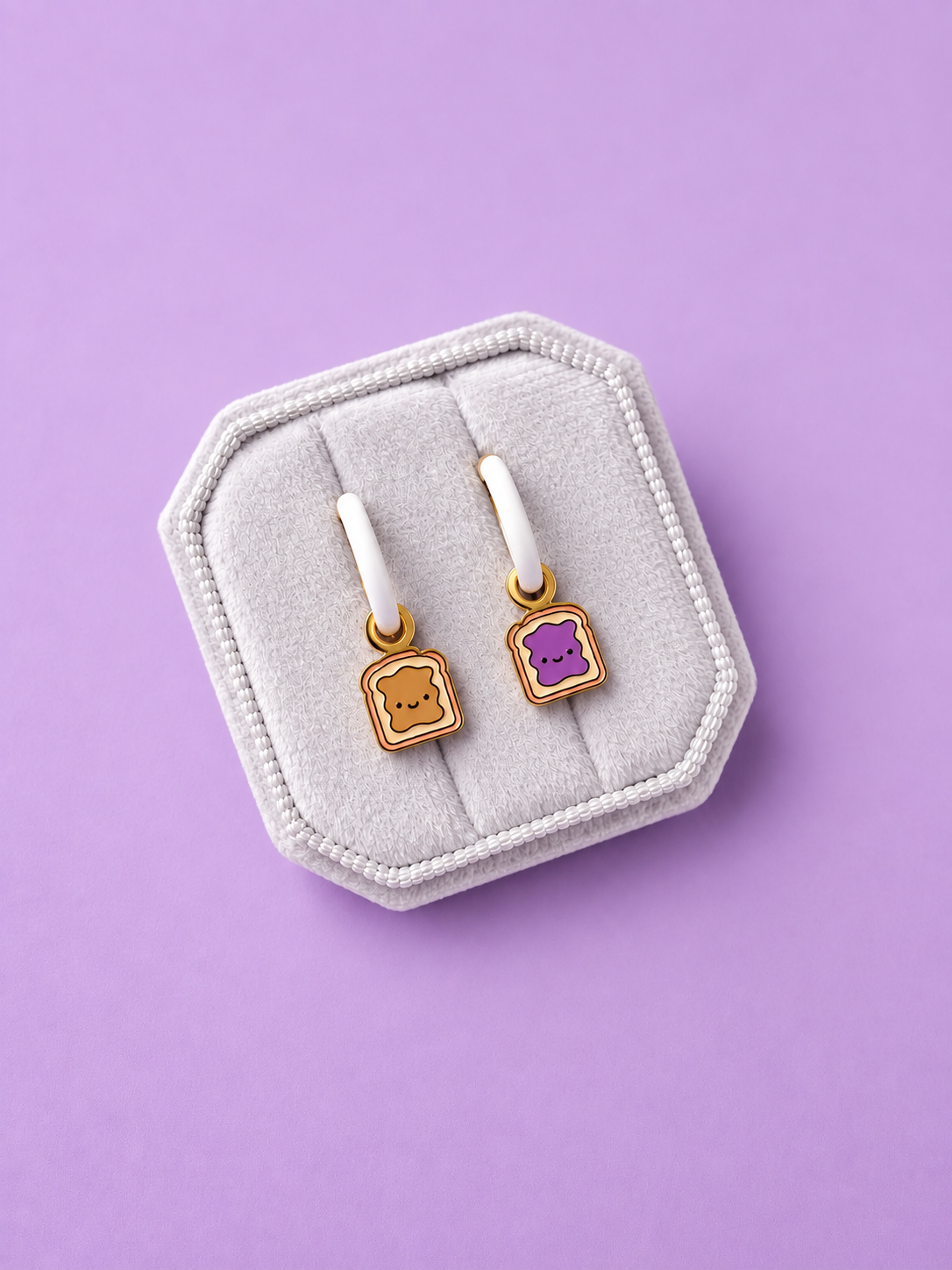A pair of gold-toned hoop earrings with small enamel charms shaped like peanut butter and jelly slices, displayed on a light gray jewelry stand against a solid lavender background.