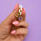 A hand with pink manicured nails holds a small jewelry charm featuring two white hoops, gold-tone metal rings, and two small peanut butter and jelly shaped charms against a solid lavender background.