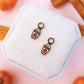 Pair of earrings on a hexagonal white stand with pumpkins and cinnamon sticks in the background.