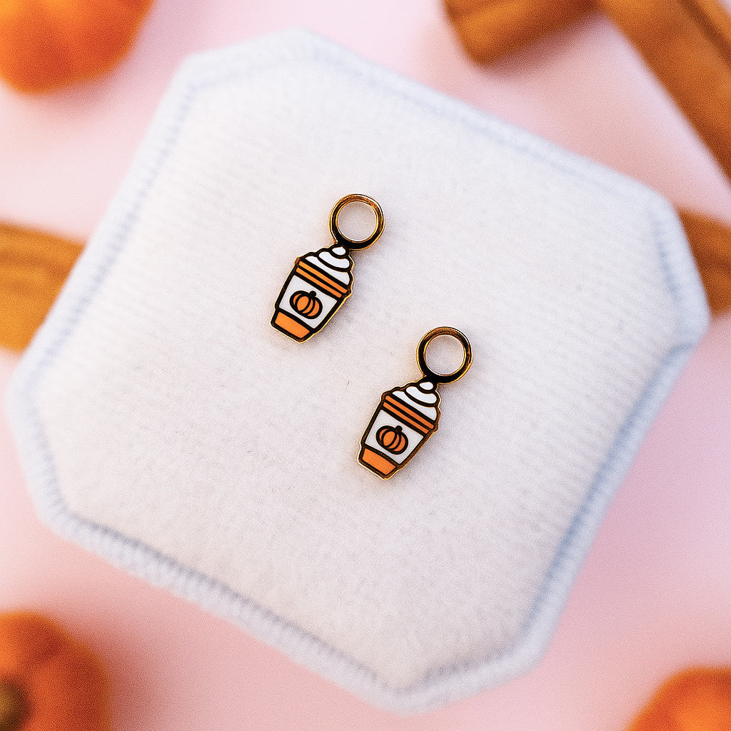 Pumpkin spice latte huggie charms on a white jewelry cushion with pumpkins and cinnamon sticks in a light pink background