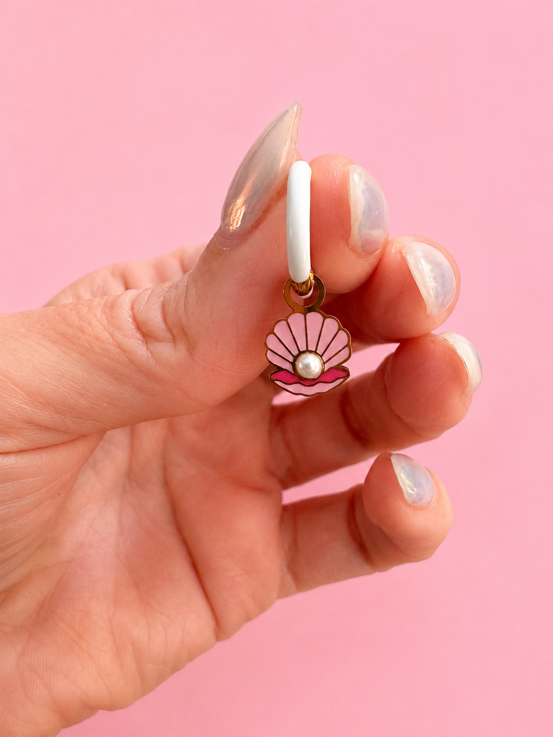 Hand holding a seashell-shaped earring with a pearl on a pink background