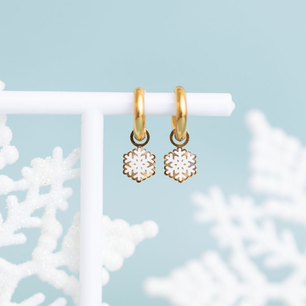 Glitter snowflake huggie charms on 8mm gold huggie hoops hanging on a white jewelry stand against a teal background with snowflakes