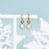 Glitter snowflake huggie charms on 8mm gold huggie hoops hanging on a white jewelry stand against a teal background with snowflakes
