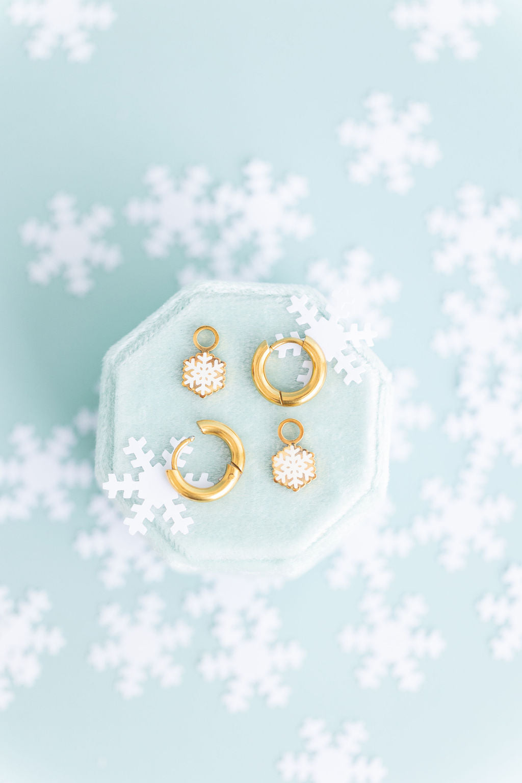 Glitter snowflake huggie charms and 8mm gold huggie hoops laying on a teal jewelry cushion on a teal background with snowflakes