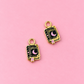 Two gold-tone book charms with black enamel detailing, featuring a crescent moon, the word 'SPELLS', and a small pink bookmark, displayed on a solid pink background.