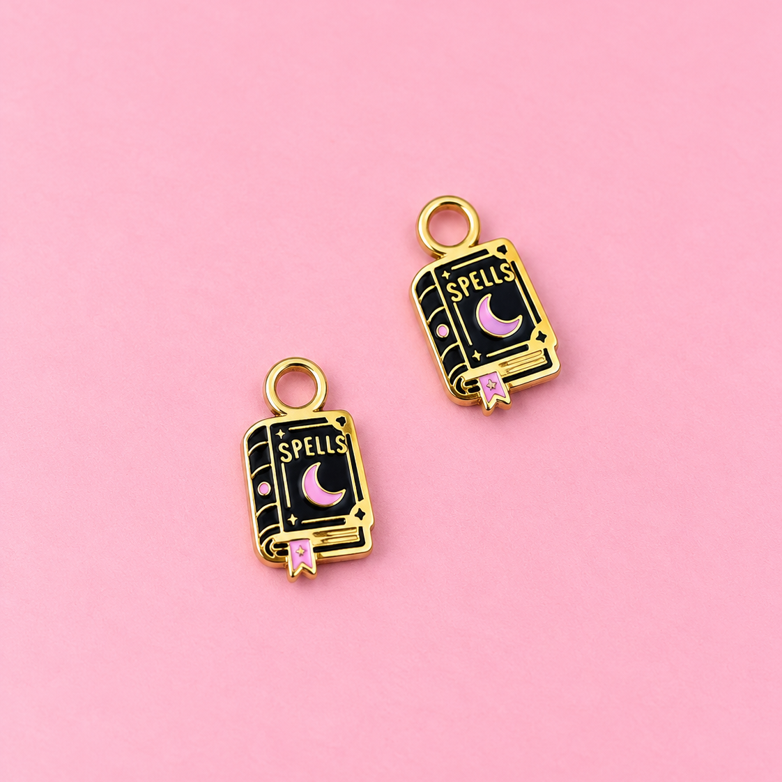 Two gold-tone book charms with black enamel detailing, featuring a crescent moon, the word 'SPELLS', and a small pink bookmark, displayed on a solid pink background.
