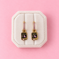 A pair of gold book-shaped drop earrings with pink rhinestone accents and a black center featuring a pink crescent moon and the word 'SPELLS', displayed on a cream-colored jewelry stand against a solid pink background.