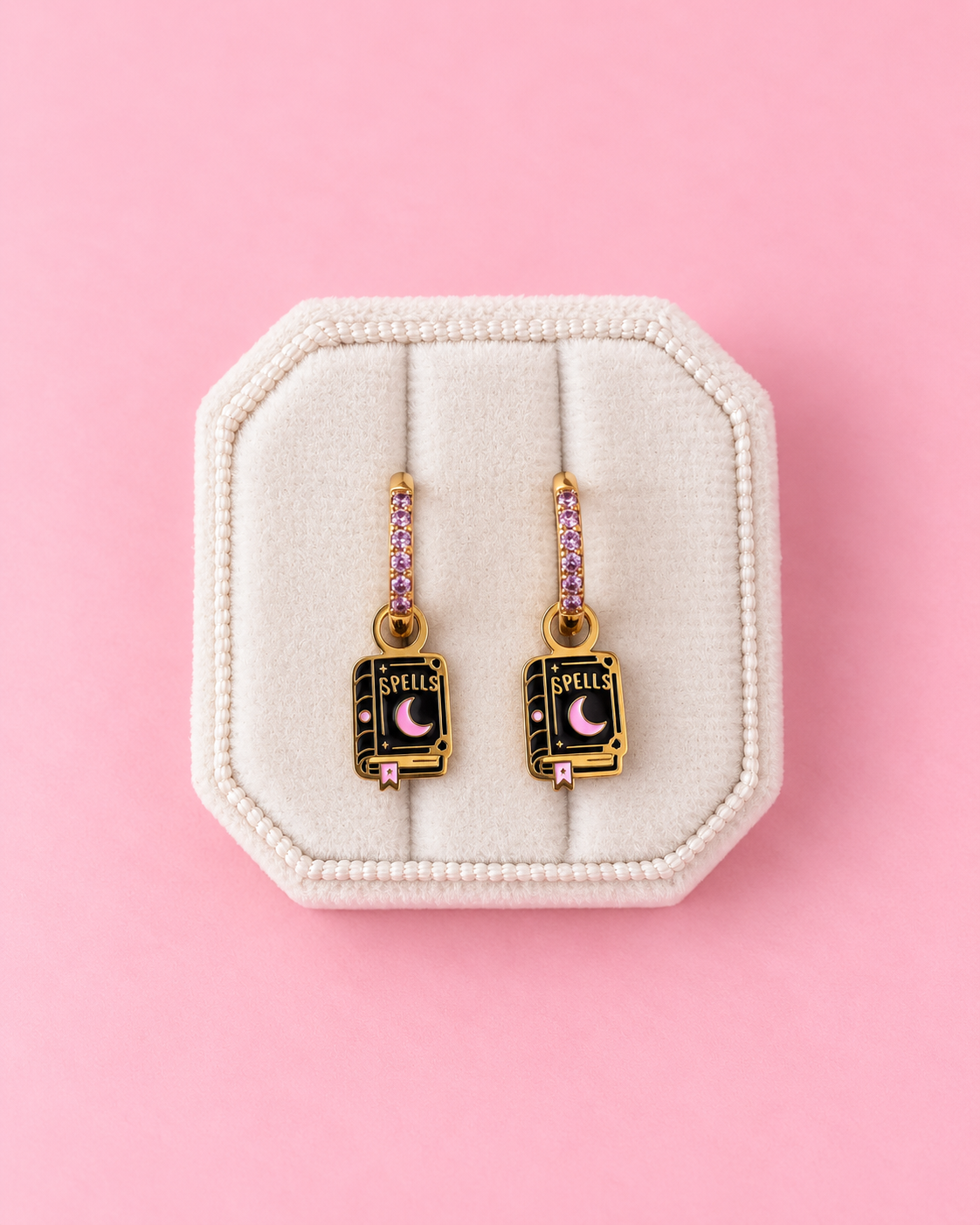 A pair of gold book-shaped drop earrings with pink rhinestone accents and a black center featuring a pink crescent moon and the word 'SPELLS', displayed on a cream-colored jewelry stand against a solid pink background.