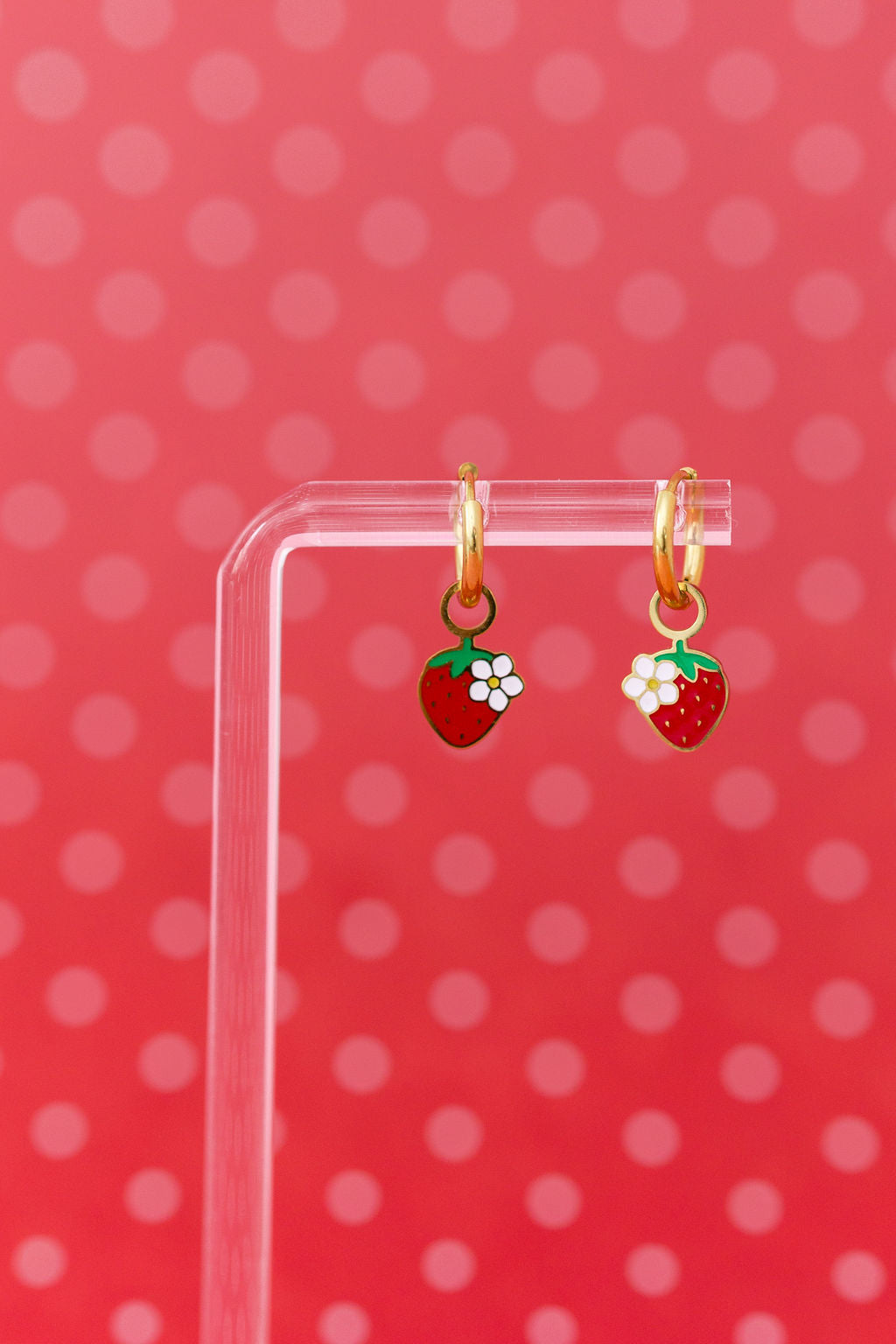 Strawberry blossom huggie charms on 10mm gold huggie hoops hanging on a clear jewelry stand against a red backdrop with pink polka dots