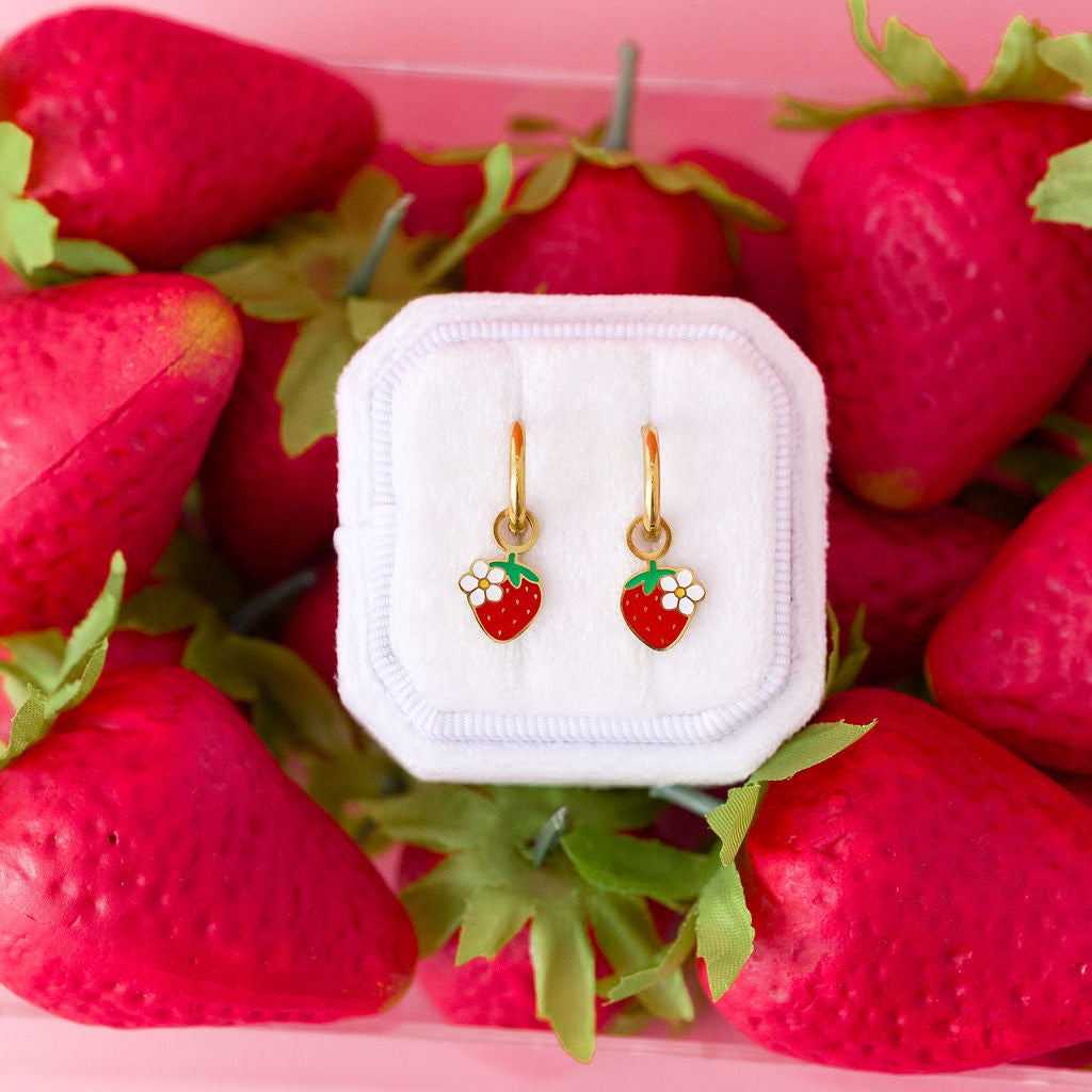 Strawberry blossom huggie charms on 10mm gold huggie hoops in a white jewelry box on top of a box of strawberries