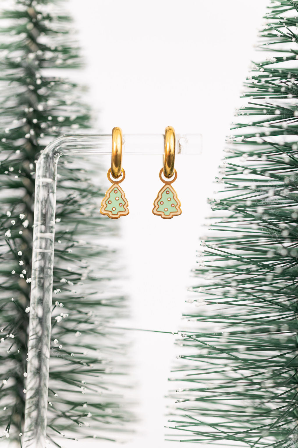 Christmas tree sugar cookie huggie charms on 8mm gold huggie hoops hanging on a clear jewelry stand against a white background with mini scrub brush Christmas trees in the background 