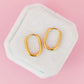 A pair of gold hoop earrings displayed on a white hexagonal jewelry stand against a solid pink background.