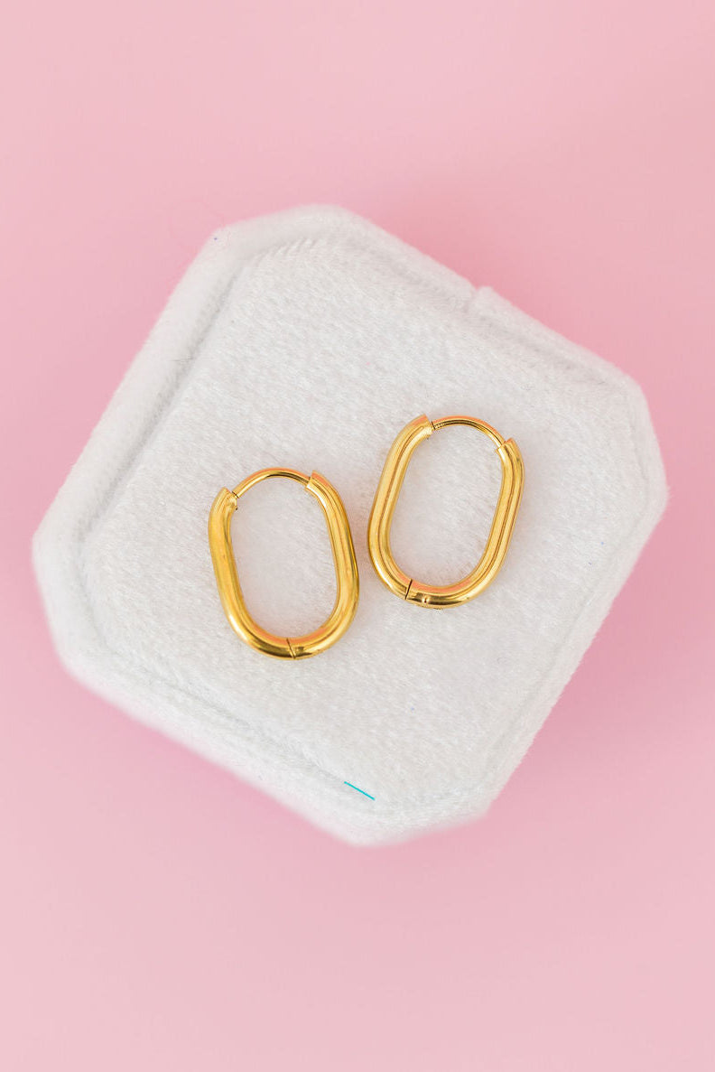 A pair of gold hoop earrings displayed on a white hexagonal jewelry stand against a solid pink background.