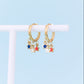 A pair of gold huggie hoop earrings with dangling star charms in blue, red, and white, displayed on a white T-stand against a light blue background.