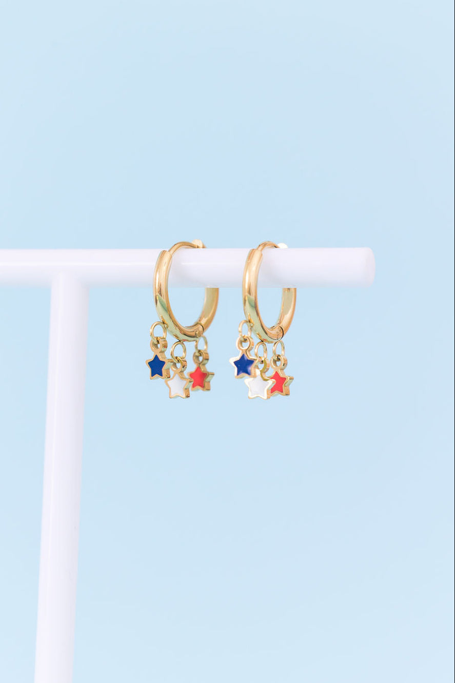 A pair of gold huggie hoop earrings with dangling star charms in blue, red, and white, displayed on a white T-stand against a light blue background.