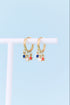 A pair of gold huggie hoop earrings with dangling star charms in blue, red, and white, displayed on a white T-stand against a light blue background.