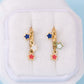 A pair of gold star-shaped dangle earrings with blue, white, and red enamel accents, displayed on a white jewelry box against a light blue background.
