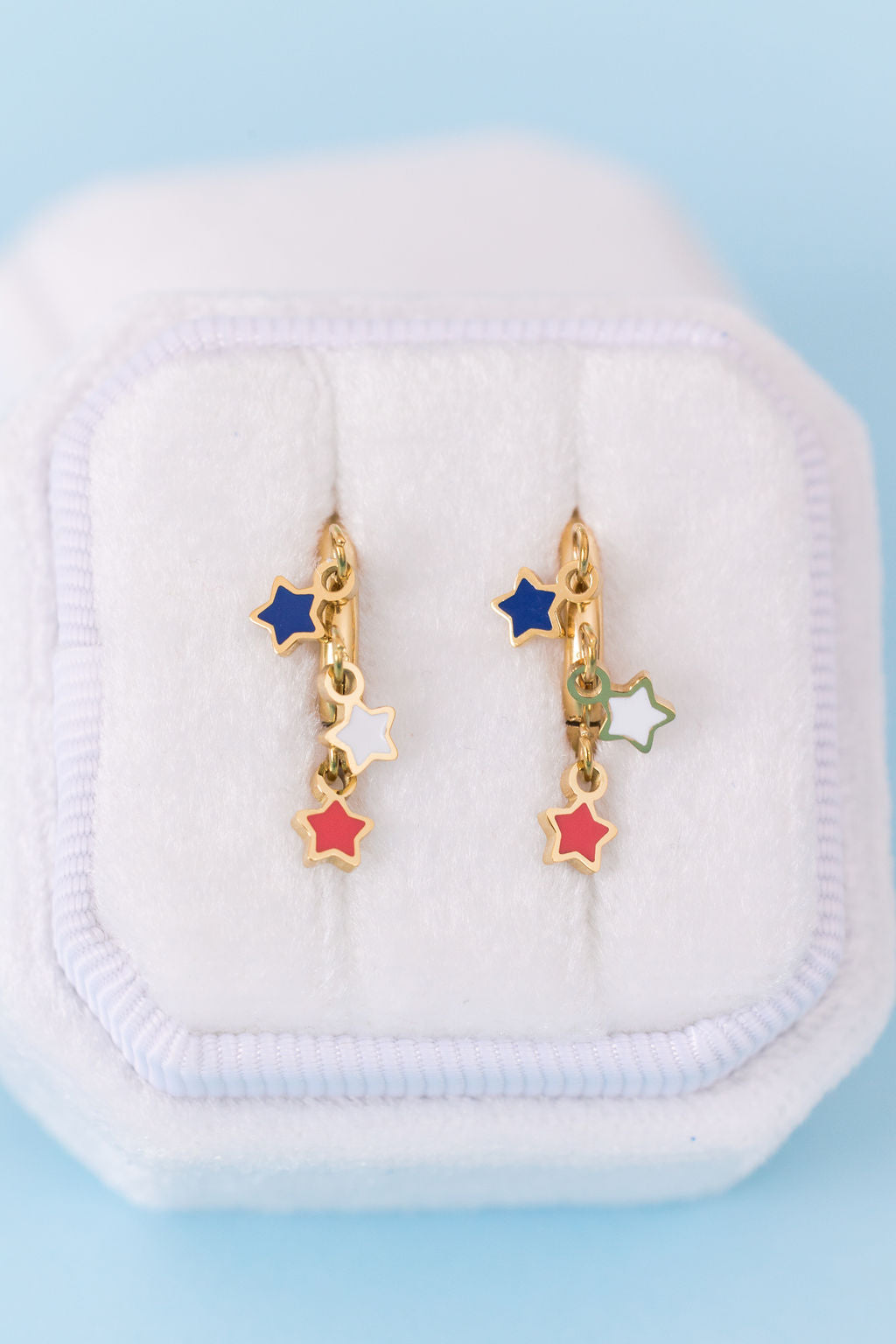 A pair of gold star-shaped dangle earrings with blue, white, and red enamel accents, displayed on a white jewelry box against a light blue background.