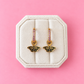A pair of gold butterfly drop earrings with pink gemstone accents, displayed on a cream-colored octagonal jewelry box against a solid pink background.