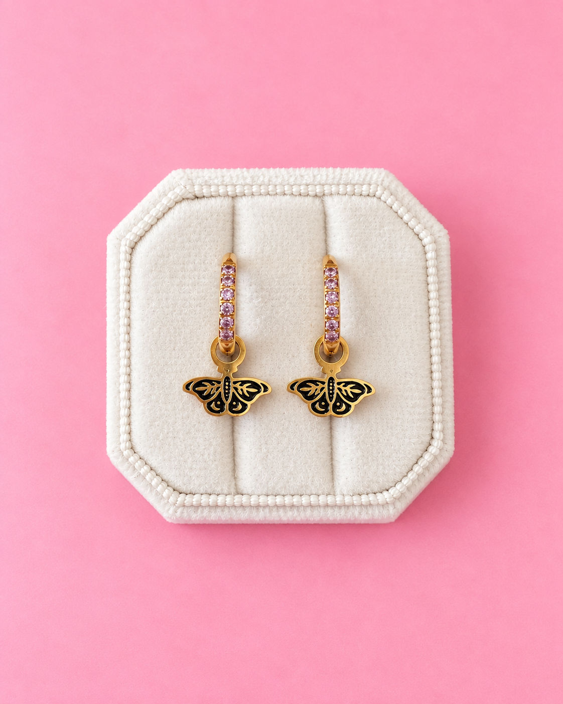 A pair of gold butterfly drop earrings with pink gemstone accents, displayed on a cream-colored octagonal jewelry box against a solid pink background.