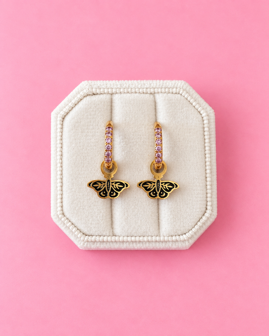 A pair of gold butterfly drop earrings with pink gemstone accents, displayed on a cream-colored octagonal jewelry box against a solid pink background.