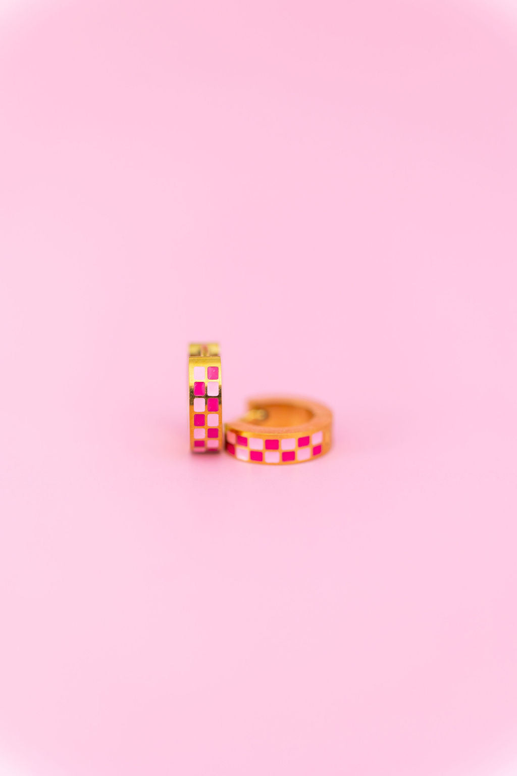 Two pink checkered chunky huggie hoops on a light pink background