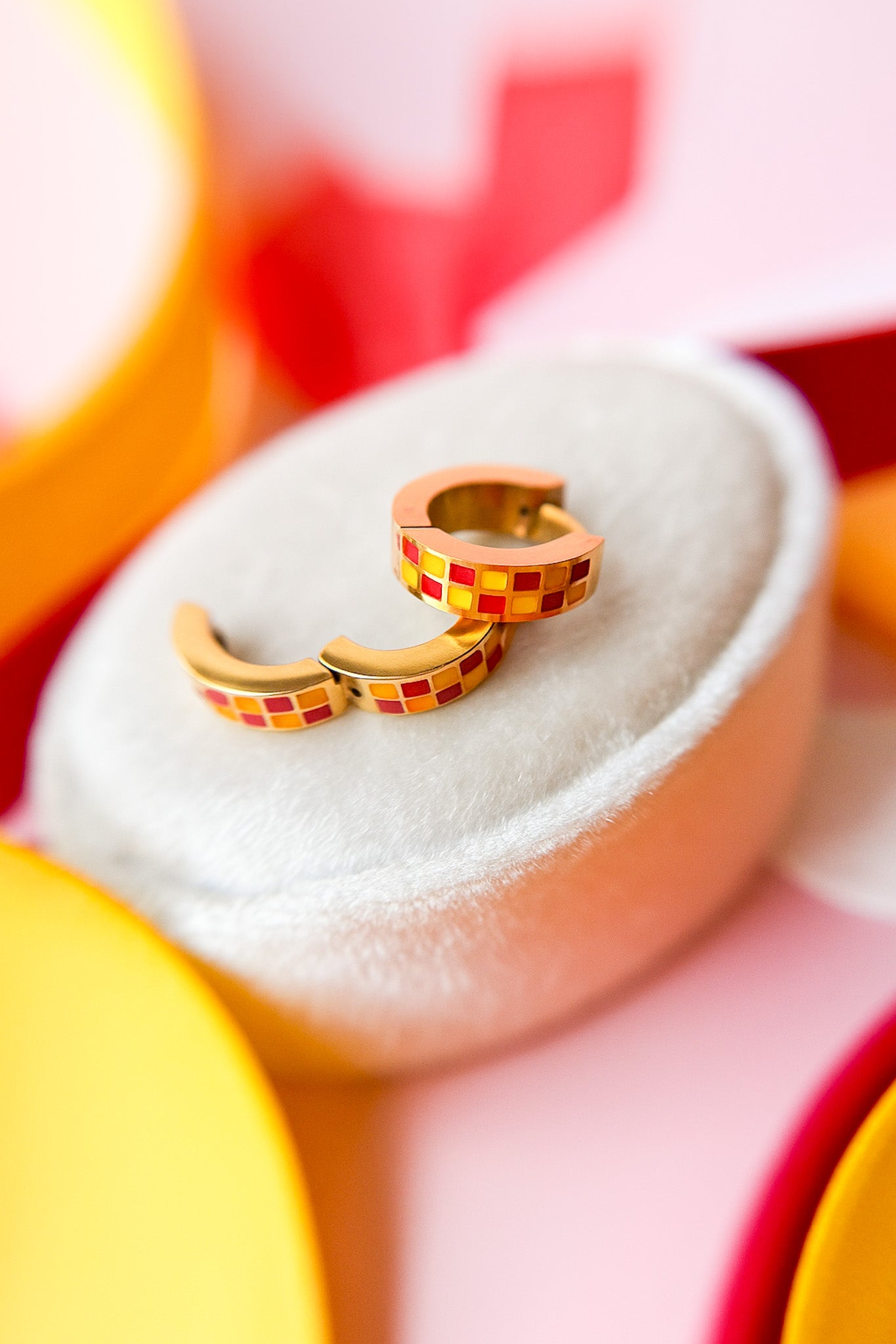 Pair of gold hoop earrings with red and yellow checkered pattern on a white cushion against a multicolored background