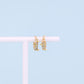 A pair of small gold hoop earrings with dangling cowboy boot charms, displayed on a white stand against a light blue background.