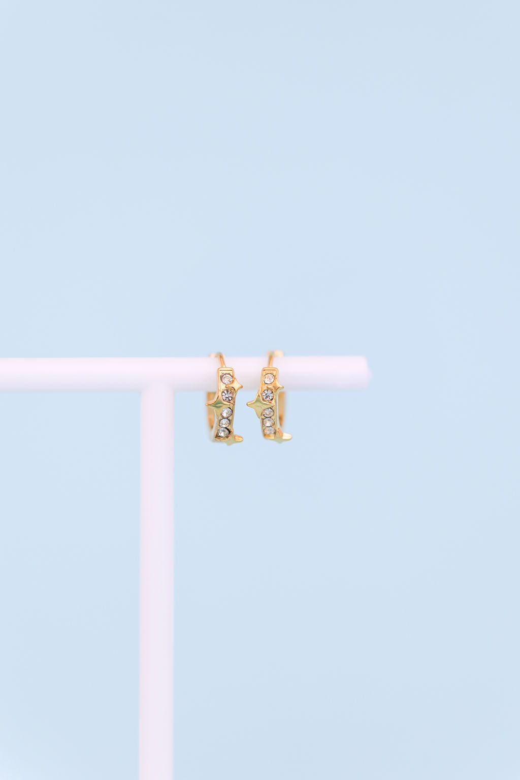 A pair of small gold hoop earrings with dangling cowboy boot charms, displayed on a white stand against a light blue background.