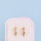 A pair of small gold huggie hoop earrings with clear rhinestones, displayed on a white velvet jewelry box against a light blue background.