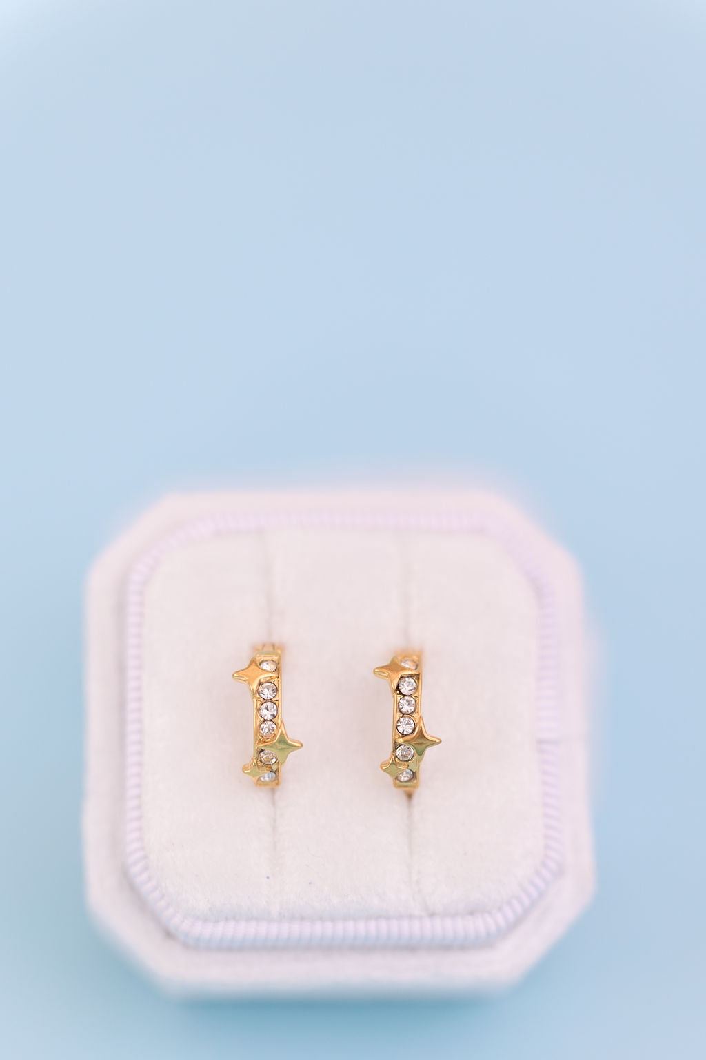 A pair of small gold huggie hoop earrings with clear rhinestones, displayed on a white velvet jewelry box against a light blue background.