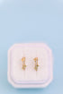 A pair of small gold huggie hoop earrings with clear rhinestones, displayed on a white velvet jewelry box against a light blue background.