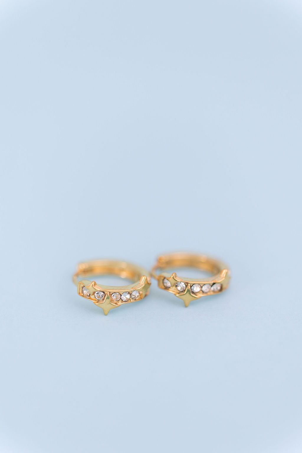 A pair of gold hoop rings with small star-shaped spikes and clear rhinestone accents, displayed on a soft light blue background.