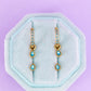 Pair of gold and turquoise earrings on a light blue hexagonal stand against a purple background