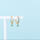 Gold hoop earrings with green gemstones on a white stand against a light blue background