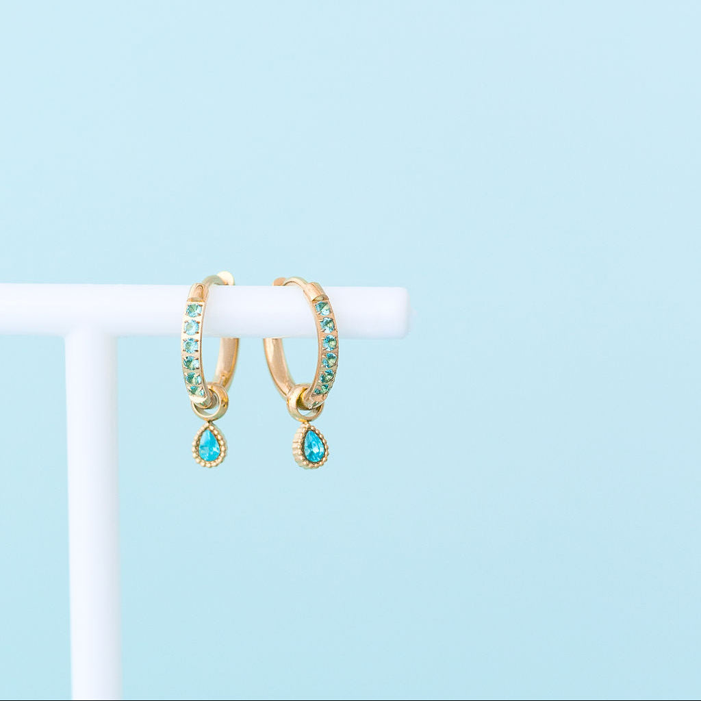 Gold hoop earrings with green gemstones on a white stand against a light blue background
