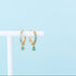 Gold hoop earrings with green gemstones on a white stand against a light blue background
