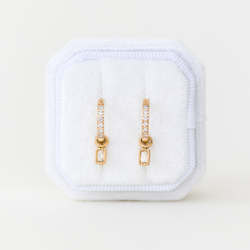 Gold cz stone huggie hoops with an emerald shape cz charm in a white jewelry box on a light beige background