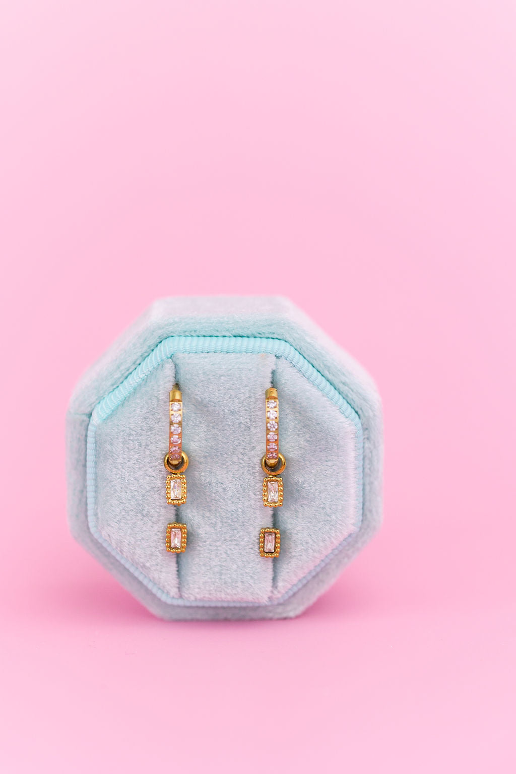 CZ Huggie hoops with emerald shaped charms and emerald shaped studs on a teal jewelry box against a light pink background