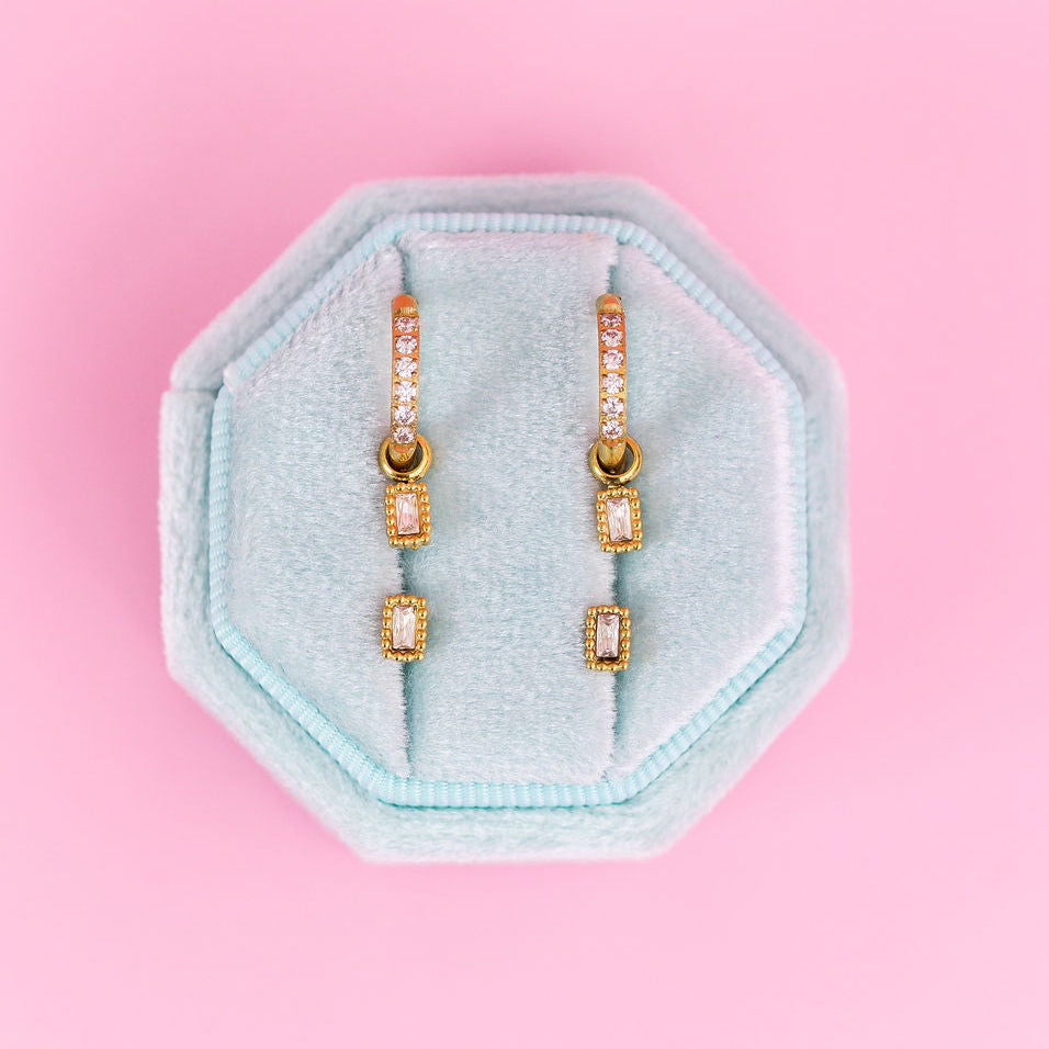 CZ Huggie hoops with emerald shaped charms and emerald shaped studs on a teal jewelry box on a light pink background
