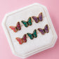 Set of three butterfly enamel studs in pink, purple and teal in a white jewelry box on a light pink background
